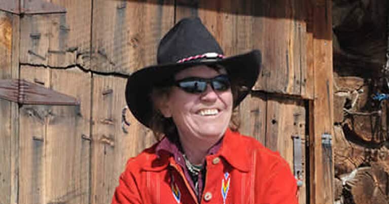 Polly grins wearing a black cowboy hat and red shirt with bright embroidery as she takes the roping practice bull by the horns.