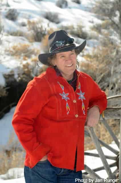 Polly Oberosler leaning on an old wagon wheel in a red shirt, black hat, and sunglasses with snowy sagebrush in the background.